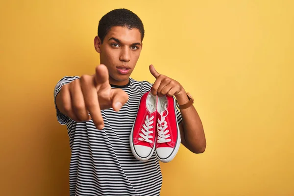 年轻的英俊阿拉伯男子拿着休闲运动鞋站在孤立的黄色背景 用手指指着相机和你 手标志 积极和自信的姿态从前面 — 图库照片