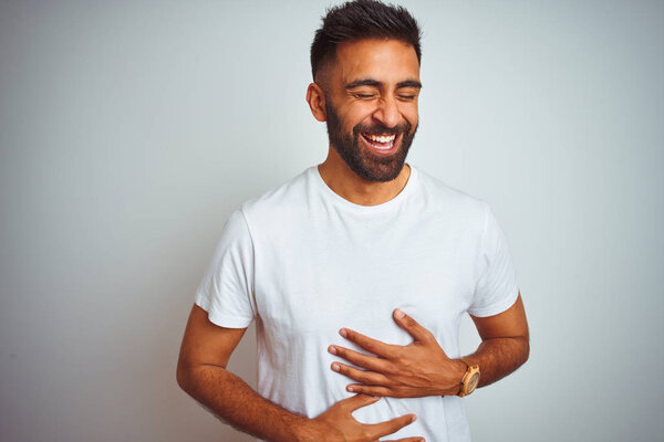 Young indian man wearing t-shirt standing over isolated white background smiling and laughing hard out loud because funny crazy joke with hands on body.
