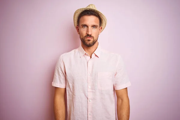 Handsome man wearing hat and summer shirt over pink isolated background with serious expression on face. Simple and natural looking at the camera.