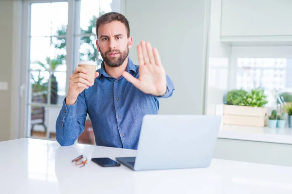 Handsome man working using computer laptop and drinking a cup of coffee with open hand doing stop sign with serious and confident expression, defense gesture
