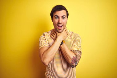 Dövme ile genç yakışıklı adam izole sarı arka plan üzerinde çizgili t-shirt giyen bağırarak ve boğulmak çünkü acı boğmak. Sağlık sorunu. Boğulma ve intihar kavramı.
