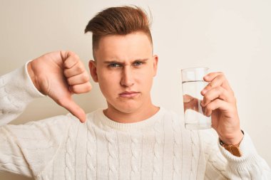 Genç yakışıklı adam kızgın yüz ile izole beyaz arka plan üzerinde duran su bardak içme, başparmak aşağı, ret kavramı ile sevmediğimi gösteren negatif işareti