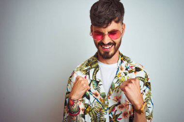 Tatilde yazlık gömlek güneş gözlüğü takan adam izole beyaz arka plan üzerinde çok mutlu ve heyecanlı kolları kaldırdı ile kazanan jest yapıyor, gülümseyerek ve başarı için çığlık. Kutlama konsepti.