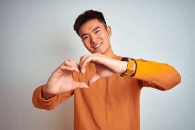 Turuncu kazak giyen genç Asyalı çinli adam izole beyaz arka plan üzerinde duran sevgi kalp sembolü ve elleriyle şekil gösteren gülümseyerek. Romantik kavram.