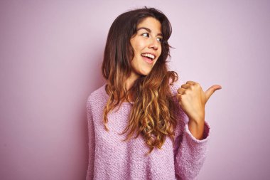 Genç güzel kadın pembe izole arka plan üzerinde duran kazak giyen mutlu yüz görünümlü gülümseyerek ve başparmak ile yan işaret.