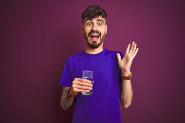 Dövme içme cam su ile izole mor arka plan üzerinde duran çok mutlu ve heyecanlı, kazanan ifade büyük bir gülümseme ile çığlık zafer kutluyor ve ellerini kaldırdı