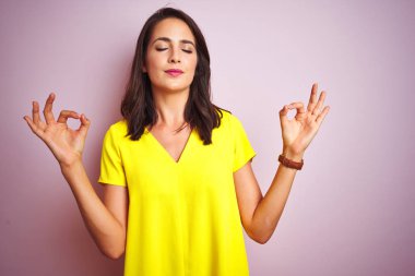 Pembe izole arka plan üzerinde duran sarı t-shirt giyen genç güzel kadın rahatlayın ve parmakları ile meditasyon jest yapıyor kapalı gözleri ile gülümseyerek. Yoga konsepti.