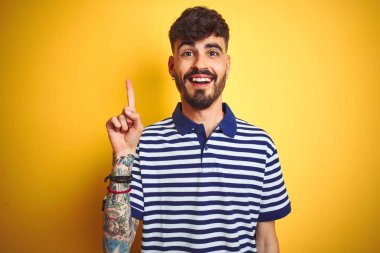 Çizgili polo giyen dövmeli genç adam izole sarı arka plan üzerinde duran başarılı bir fikir ile parmak işaret. Çıktı ve mutlu. Bir numara.