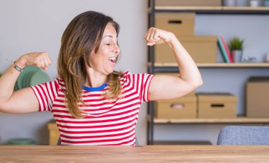Orta yaş yaşlı kadın evde masada oturan kol kasları gururla gülümseyerek gösteren. Fitness konsepti.