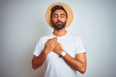 Tatilde yazlık şapka giyen genç hintli adam izole beyaz arka plan üzerinde kapalı gözleri ve yüzünde minnettar jest ile göğüs üzerinde gülümseyerek ayakta. Sağlık kavramı.