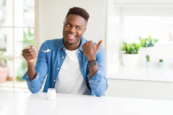 African american man eating healthy natural yogurt with a spoon pointing and showing with thumb up to the side with happy face smiling
