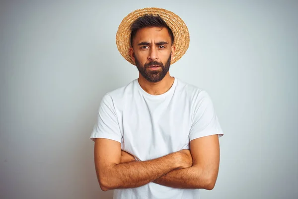 Young indian man on holiday wearing summer hat standing over isolated white background skeptic and nervous, disapproving expression on face with crossed arms. Negative person.