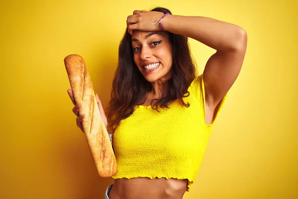 Young beautiful baker woman holding bread standing over isolated yellow background stressed with hand on head, shocked with shame and surprise face, angry and frustrated. Fear and upset for mistake.