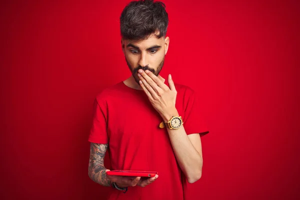 Young man with tattoo using tablet standing over isolated red background cover mouth with hand shocked with shame for mistake, expression of fear, scared in silence, secret concept