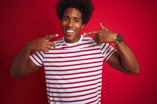孤立した赤い背景の上にストライプのTシャツを着たアフロヘアの若いアフリカ系アメリカ人男性は 明るく見せ 指の歯と口で指を向けます 歯科健康の概念 — ストック写真