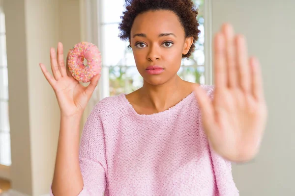 Young african american woman eating pink sugar donut with open hand doing stop sign with serious and confident expression, defense gesture