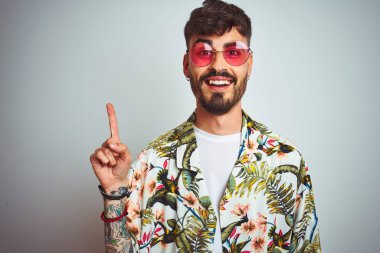 Tatilde dövmeli adam başarılı bir fikir ile parmak işaret izole beyaz arka plan üzerinde yazlık gömlek güneş gözlüğü giyiyor. Çıktı ve mutlu. Bir numara.