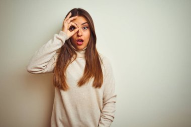 İzole beyaz arka plan üzerinde kış boğazlı kazak giyen genç güzel kadın ok jest yaparak şaşırmış yüz, göz parmakları ile bakarak şok. İnanmayan ifade.