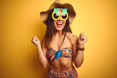 Bikini ve ananas güneş gözlüğü takan tatilde izole sarı arka plan üzerinde kutlayan kadın şaşırmış ve kollarını kaldırdı ve gözleri açık başarı için şaşırttı. Kazanan kavramı.