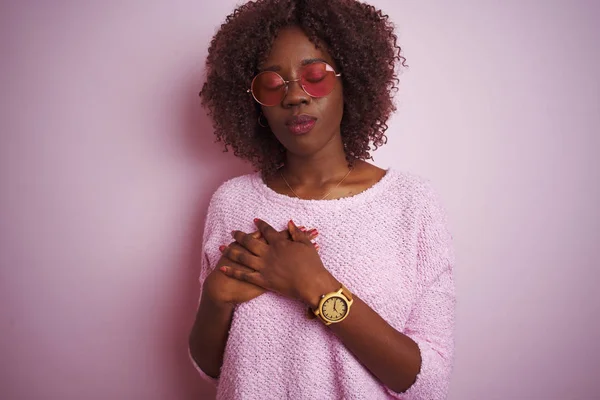 Young african afro woman wearing sweater and sunglasses over isolated pink background smiling with hands on chest with closed eyes and grateful gesture on face. Health concept.