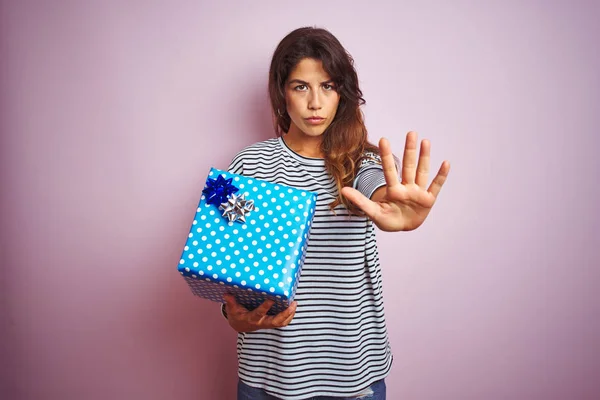 Young beautiful woman holding birthday gift over pink isolated background with open hand doing stop sign with serious and confident expression, defense gesture