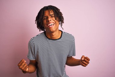 Izole pembe arka plan üzerinde duran lacivert çizgili t-shirt giyen dreadlocks ile Afro adam çok mutlu ve heyecanlı silah kaldırdı ile kazanan jest yapıyor, gülümseyen ve başarı için çığlık. Kutlama konsepti.