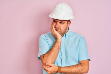 İzole edilmiş pembe arka plan üzerinde duran güvenlik kaskı takan genç mimar adam yorgun ve çapraz kolları ile depresyon sorunları ile sıkılmış görünümlü düşünme.