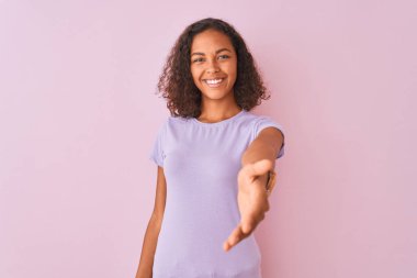 Genç Brezilyalı kadın izole pembe arka plan üzerinde duran t-shirt giyen tebrik ve karşılama olarak dostça sunan el sıkışma gülümseyerek. Başarılı iş.