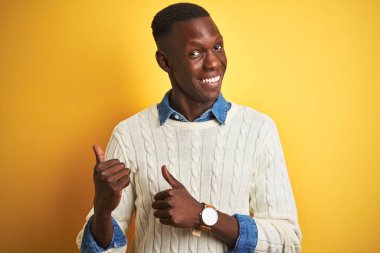 Izole sarı arka plan üzerinde denim gömlek ve beyaz kazak giyen Afro-Amerikan adam el ve başparmak yukarı arka işaret, kendinden emin gülümseyerek
