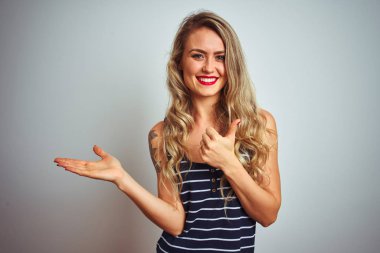 Genç güzel kadın çizgili t-shirt beyaz izole arka plan üzerinde duran giyen palmiye el gösteren ve başparmak ile ok jest yapıyor, mutlu ve neşeli gülümseyerek