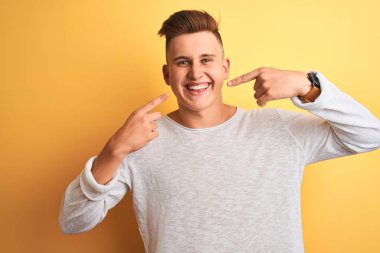 Genç yakışıklı adam izole sarı arka plan üzerinde duran beyaz rahat t-shirt giyen neşeli gösteren ve parmakları diş ve ağız ile işaret gülümseyerek. Diş sağlığı kavramı.
