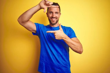 Genç yakışıklı adam sarı izole arka plan üzerinde rahat mavi t-shirt giyen mutlu yüz ile elleri ve parmakları ile çerçeve yapma gülümseyerek. Yaratıcılık ve fotoğrafçılık konsepti.