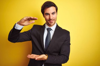 Takım elbise giyen ve kravat giyen genç yakışıklı işadamı elleri büyük ve büyük boy işareti gösteren izole sarı arka plan üzerinde duran, sembol ölçmek. Kameraya bakarak gülümsüyor. Ölçme kavramı.