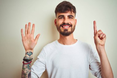 Kendine güvenen ve mutlu gülümserken izole beyaz arka plan üzerinde duran dövme giyen genç adam gösteren ve parmakları ile işaret altı.