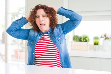 Evde kot ceket giyen kıvırcık saçlı orta yaş yaşlı kadın Deli ve baş elleri ile korkmuş, korkmuş ve açık ağız ile şok şaşırttı