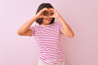 İzole pembe arka plan üzerinde duran çizgili t-shirt giyen genç güzel kadın işaret bakarak gülümseyen el ve parmakları ile kalp şekli yapıyor