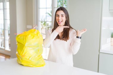 Çöp konteynerinden çöpü alan güzel genç kadın, el iyle sunarken ve parmakla işaret ederken kameraya gülümseyerek.