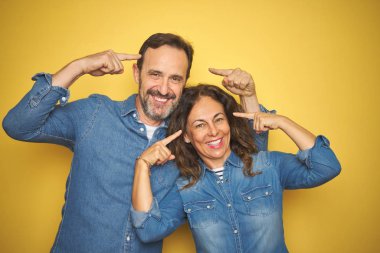 Güzel orta yaş çift birlikte izole sarı arka plan üzerinde iki elparmağı, harika bir fikir ya da düşünce, iyi bellek ile baş gösteren gülümseyerek ayakta