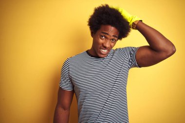 Genç afro-amerikan adam izole sarı arka plan üzerinde duran eldiven kullanarak temizlik baş üzerinde el ile vurguladı, utanç ve sürpriz yüz ile şok, kızgın ve sinirli. Korku ve hata için üzgün.