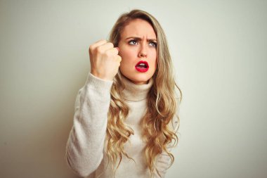 Izole beyaz arka plan üzerinde kış boğazlı kazak giyen güzel kadın kızgın ve öfkeli yükselterek yumruk sinirli ve öfkeli öfke ile bağırırken. Öfke ve agresif kavram.