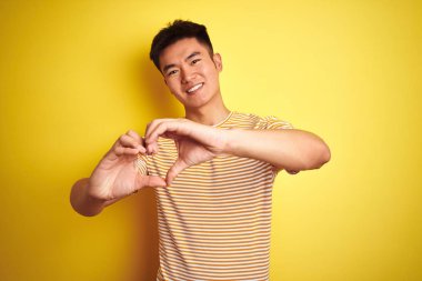 Genç Asyalı Çinli adam izole sarı arka plan üzerinde duran t-shirt giyen aşk elleri ile kalp sembolü şekli yapıyor gülümseyerek. Romantik kavram.