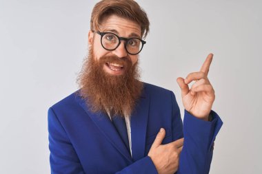 Genç kızıl İrlandalı işadamı yüzünde büyük bir gülümseme ile izole beyaz arka plan üzerinde takım elbise ve gözlük giyen, kameraya bakarak yan el ve parmak ile işaret.