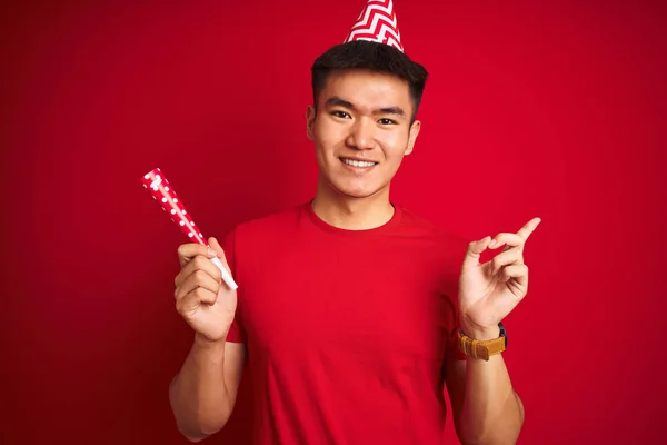 Asian chinese man on birthday celebration wearing funny hat over isolated red background very happy pointing with hand and finger to the side