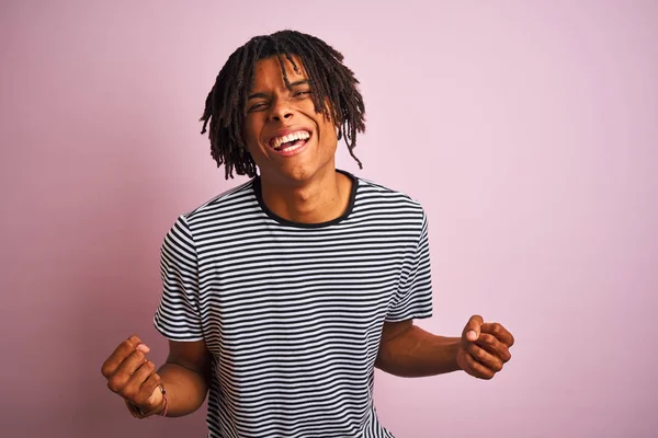 Izole pembe arka plan üzerinde duran lacivert çizgili t-shirt giyen dreadlocks ile Afro adam çok mutlu ve heyecanlı silah kaldırdı ile kazanan jest yapıyor, gülümseyen ve başarı için çığlık. Kutlama konsepti.