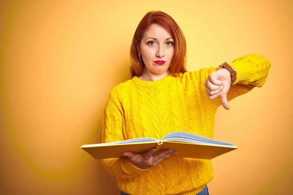 Young redhead student woman reading book standing over yellow isolated background with angry face, negative sign showing dislike with thumbs down, rejection concept
