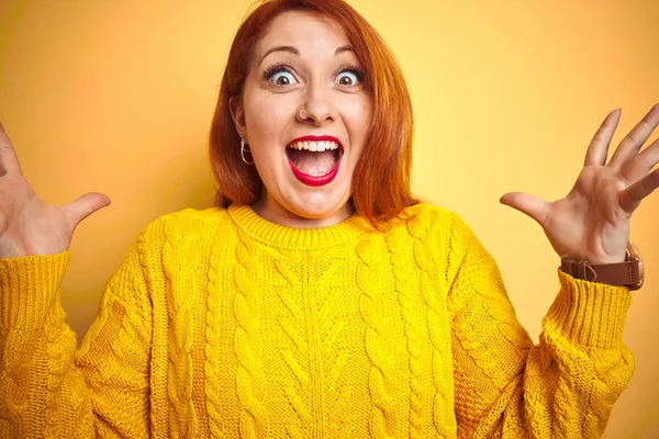Genç kızıl saçlı kadın izole sarı arka plan üzerinde duran kış kazak giyen deli kutluyor ve kollarını kaldırdı ve açık gözleri heyecan çığlık ile başarı için hayran. Kazanan kavramı