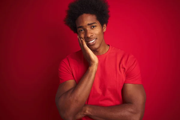 Young american man with afro hair wearing t-shirt standing over isolated red background thinking looking tired and bored with depression problems with crossed arms.