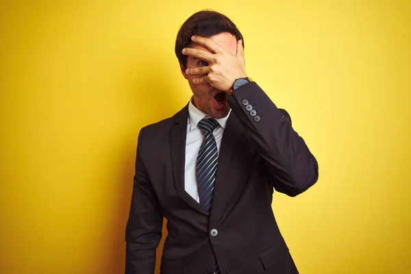 Young handsome businessman wearing suit and tie standing over isolated yellow background peeking in shock covering face and eyes with hand, looking through fingers with embarrassed expression.