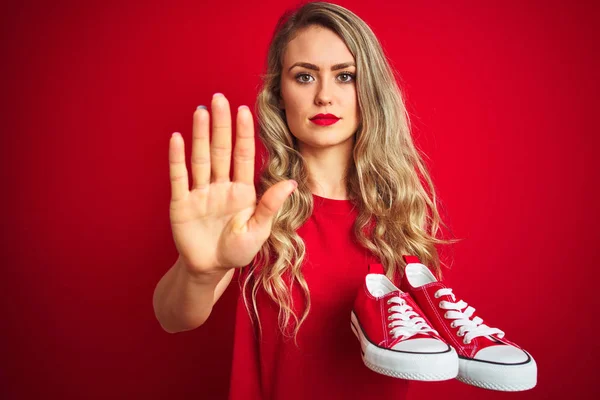 Young beautiful woman holding fashion sneakers over red isolated background with open hand doing stop sign with serious and confident expression, defense gesture