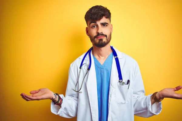 Young doctor man with tattoo wearing stethocope standing over isolated yellow background clueless and confused expression with arms and hands raised. Doubt concept.